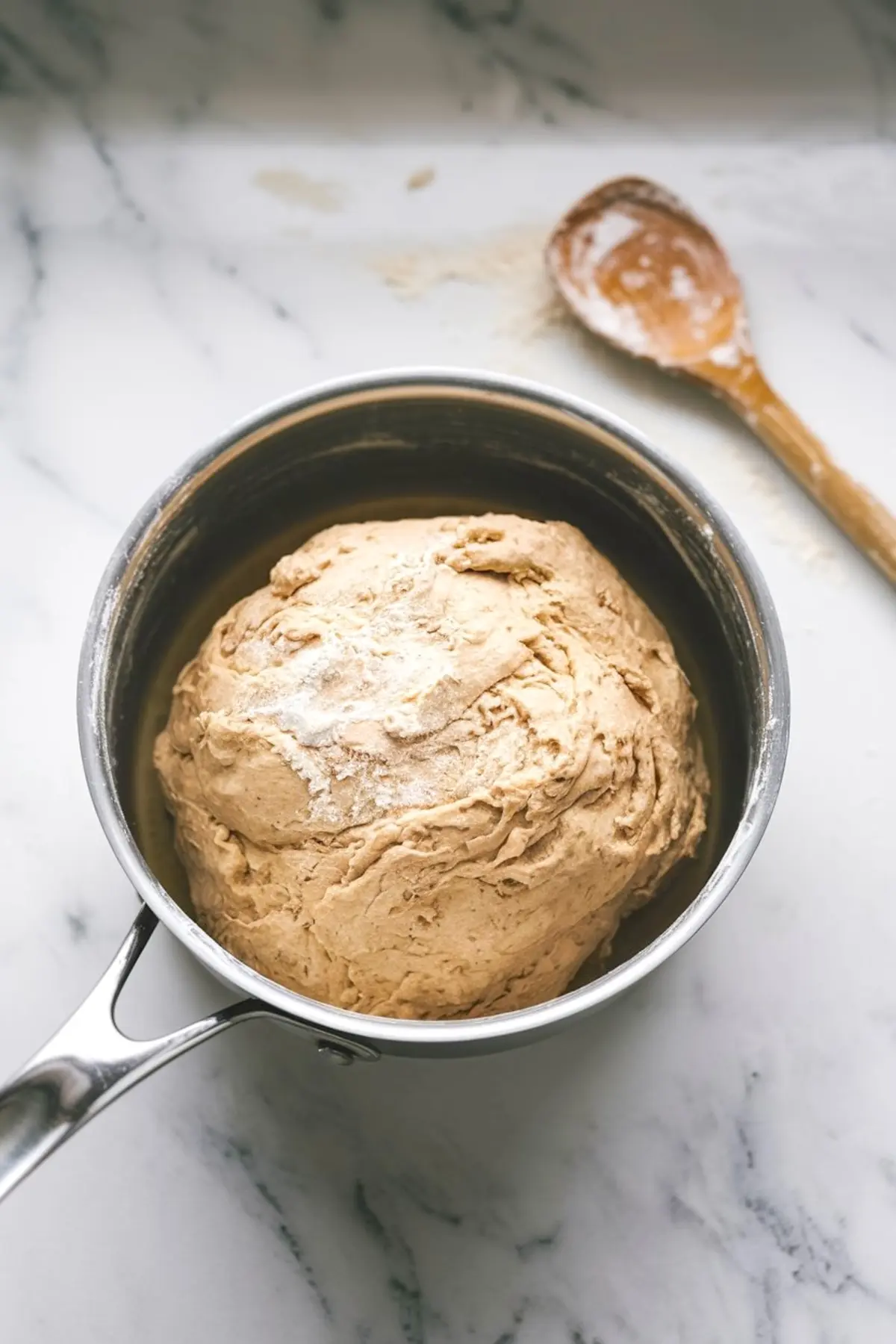 Stainless steel pot filled with freshly mixed choux pastry dough resting on a floured marble surface next to a wooden spoon.
