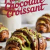 Close-up of a baked croissant drizzled with dark chocolate and vibrant green pistachio sauce, topped with crushed pistachios, with more croissants and milk jars in the background.