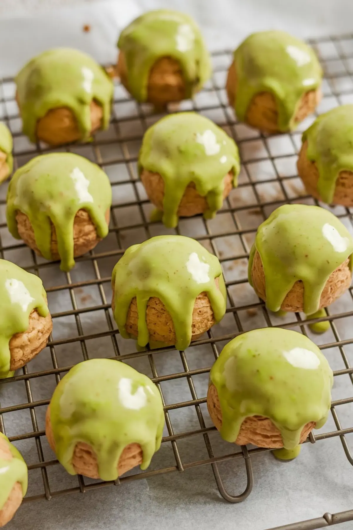 Freshly baked pistachio cream puffs coated in glossy green glaze cooling on a wire rack, arranged in neat rows over parchment-lined tray.