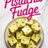 Overhead view of pistachio fudge squares arranged on a round ceramic plate. The pale green fudge is studded with whole pistachios, creating a vibrant contrast of green, yellow, and purple hues. Bold pink script reading “Pistachio Fudge” overlays the marble background.