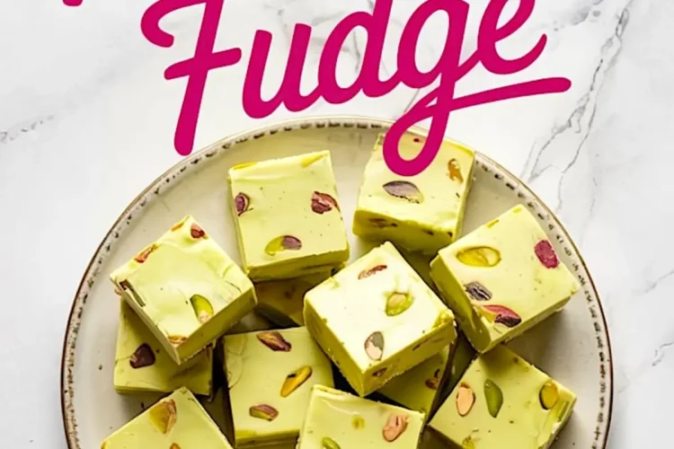 Overhead view of pistachio fudge squares arranged on a round ceramic plate. The pale green fudge is studded with whole pistachios, creating a vibrant contrast of green, yellow, and purple hues. Bold pink script reading “Pistachio Fudge” overlays the marble background.