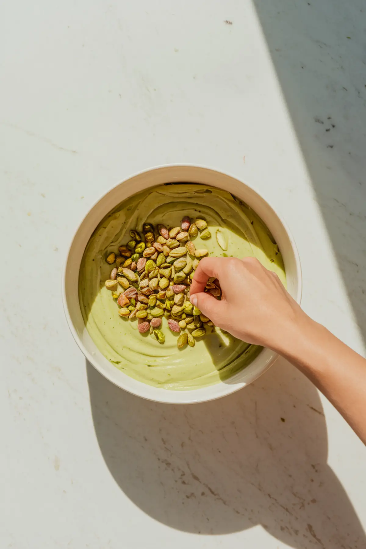 Hand sprinkling whole pistachios into a bowl of pale green pistachio fudge mixture. The creamy batter is swirled smoothly inside a white mixing bowl on a marble surface under natural light.