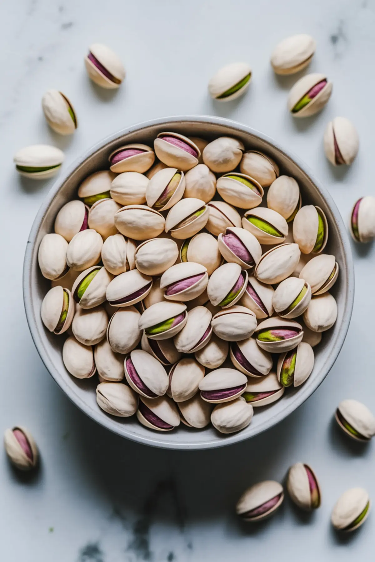 Close-up of a bowl filled with shelled whole pistachios, showcasing their natural cream, green, and purple hues, arranged on a light marble surface.