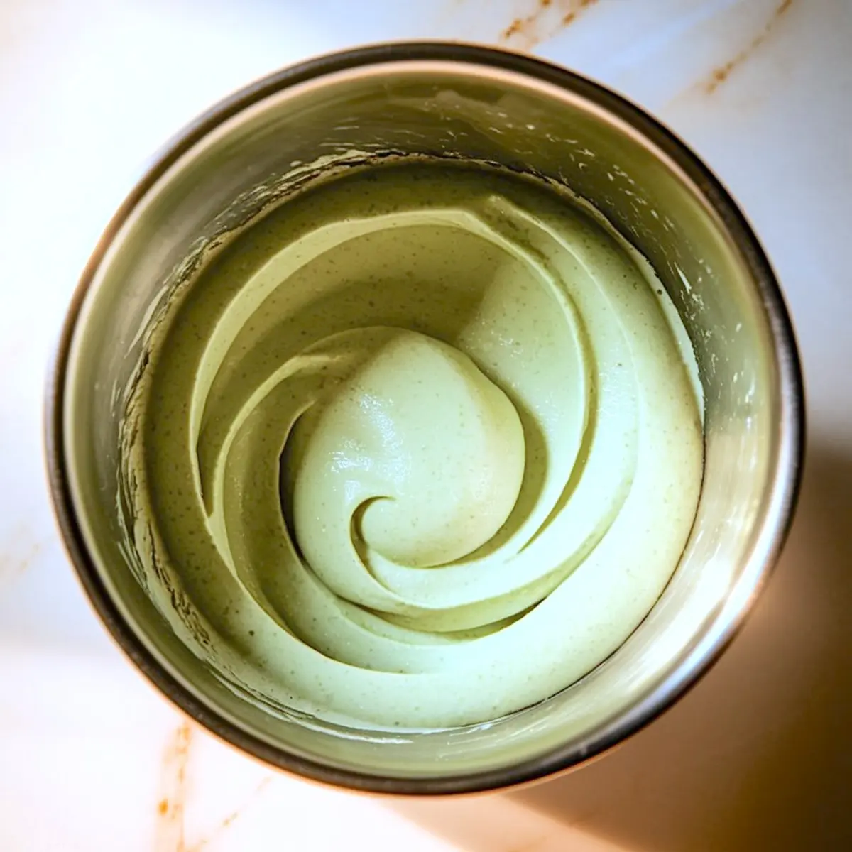 Overhead view of creamy pistachio mousse with a smooth spiral texture inside a metal mixing bowl, lit by warm lighting.
