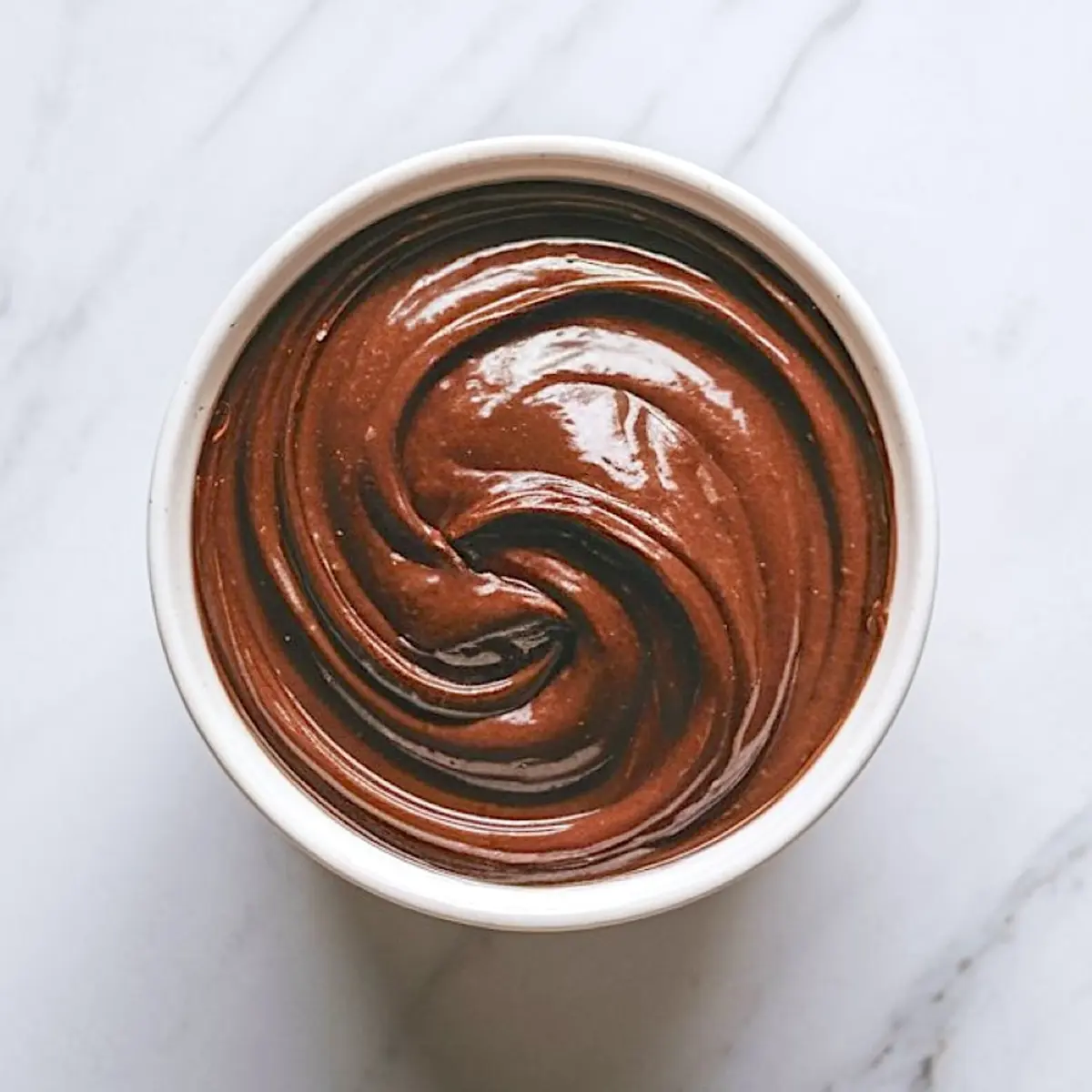 Overhead shot of glossy chocolate brownie batter swirled in a white ceramic bowl on a marble surface.
