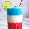 Close-up of a vibrant red, white, and blue slushie in a tall glass with a lime slice and striped paper straw, condensation on the glass highlighting the icy texture, placed on a white marble surface.