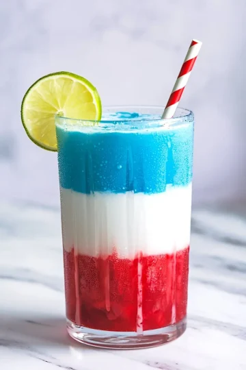 Close-up of a vibrant red, white, and blue slushie in a tall glass with a lime slice and striped paper straw, condensation on the glass highlighting the icy texture, placed on a white marble surface.