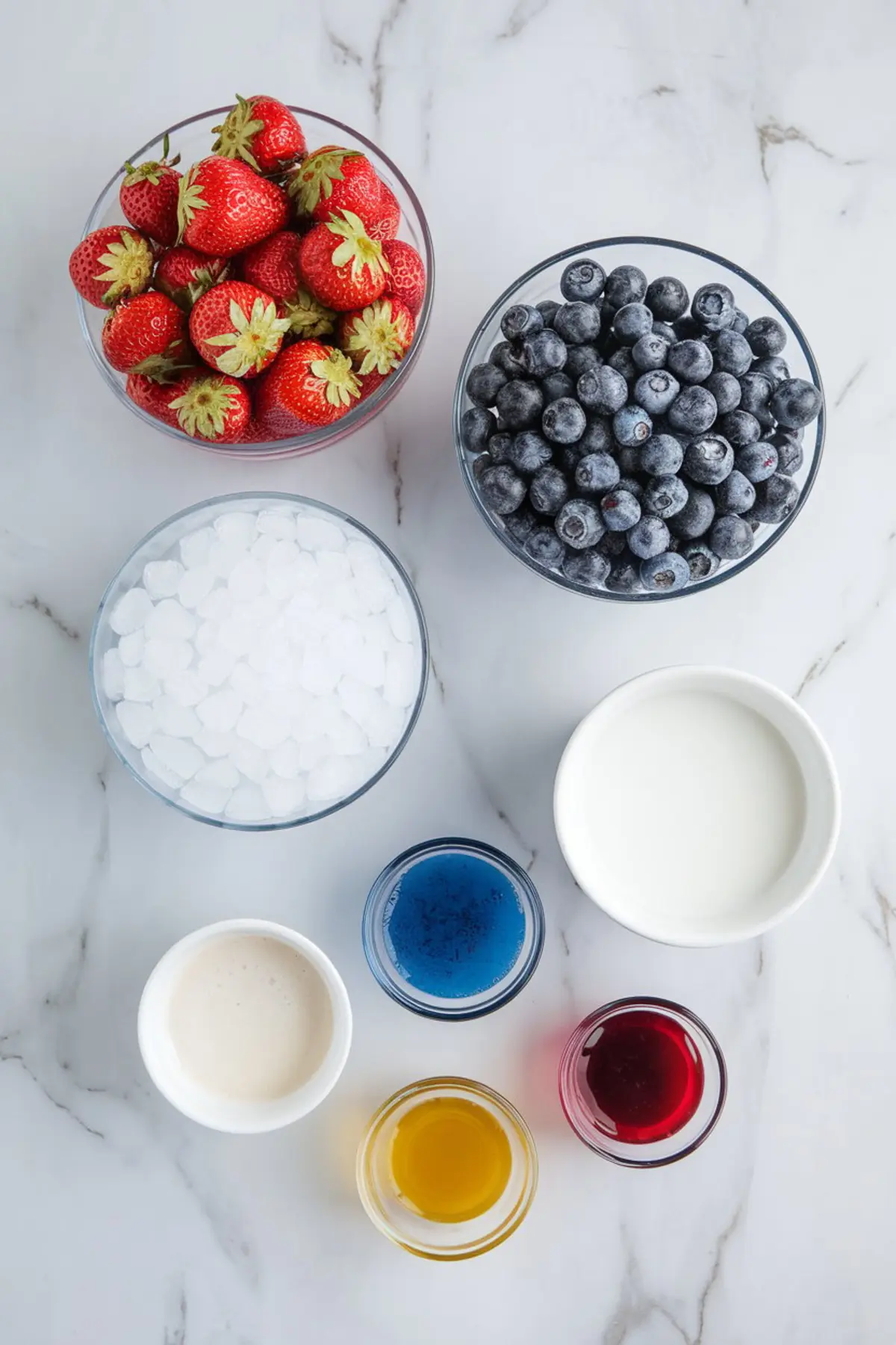 Flat lay of slushie ingredients on a marble background including fresh strawberries, blueberries, crushed ice, coconut milk, red and blue syrup, honey, and creamy bases arranged in clear bowls.
