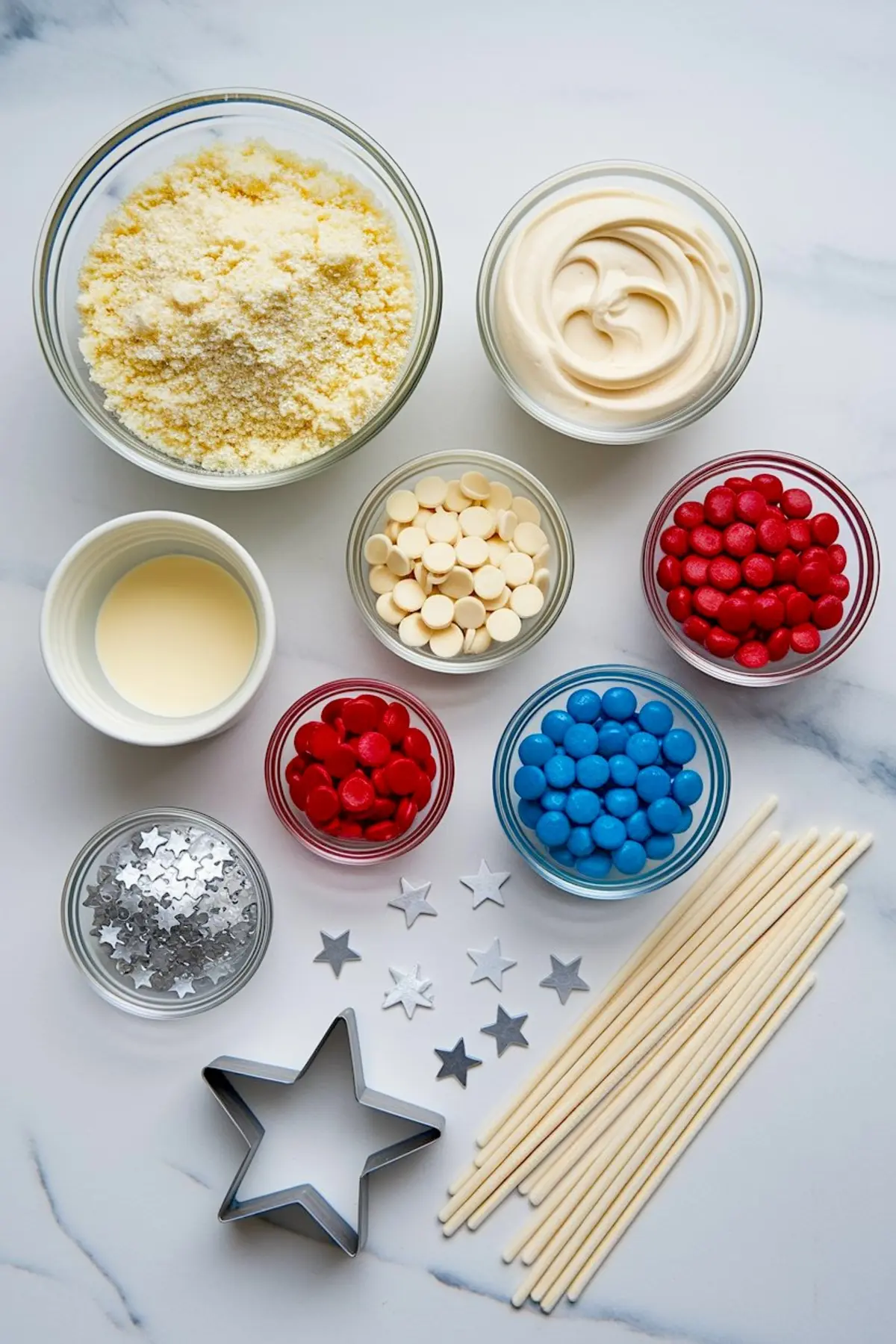 Flat lay of cake pop ingredients including crumbled cake, frosting, candy melts, red and blue candies, edible silver stars, lollipop sticks, and a star-shaped cookie cutter on a marble surface.