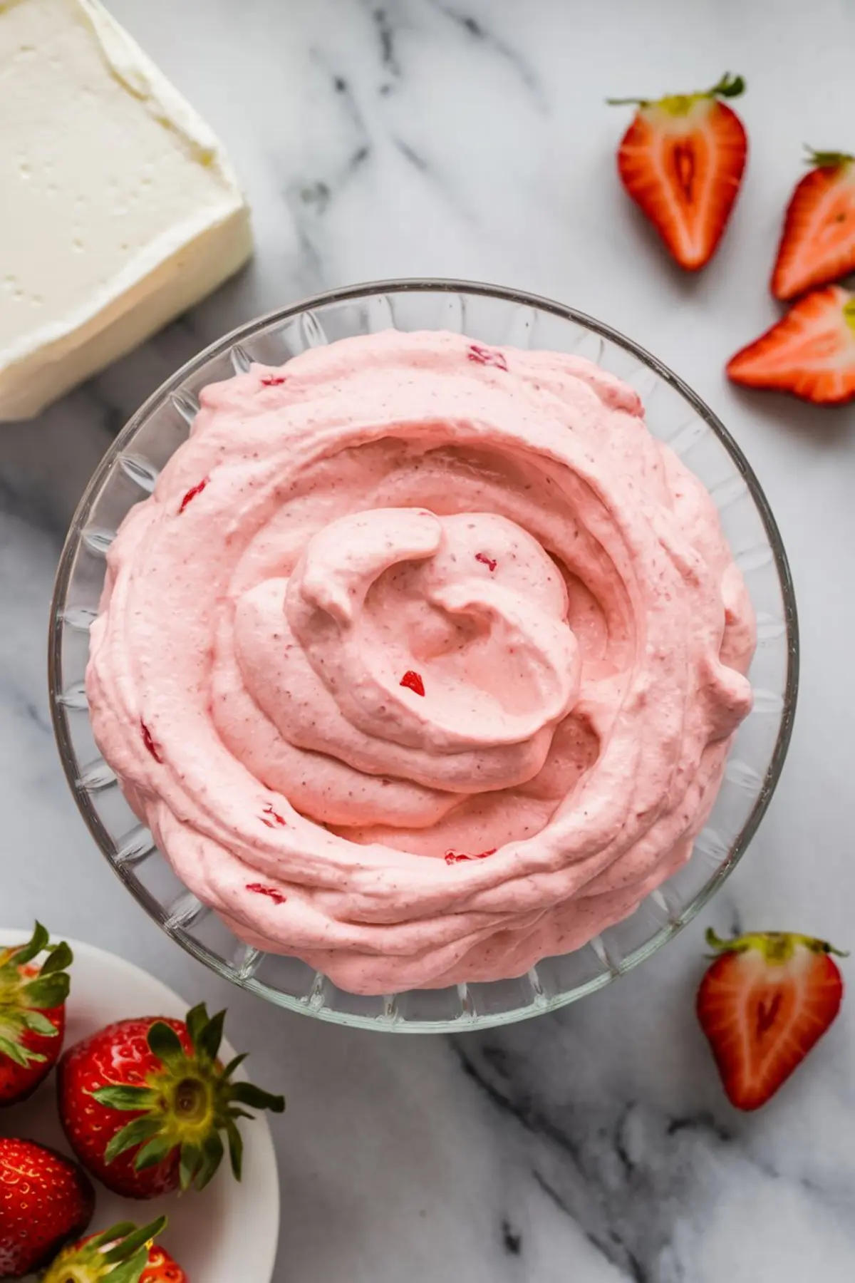 Bowl of fluffy strawberry frosting with pink cream cheese blend and visible strawberry bits, surrounded by halved strawberries on a marble surface.