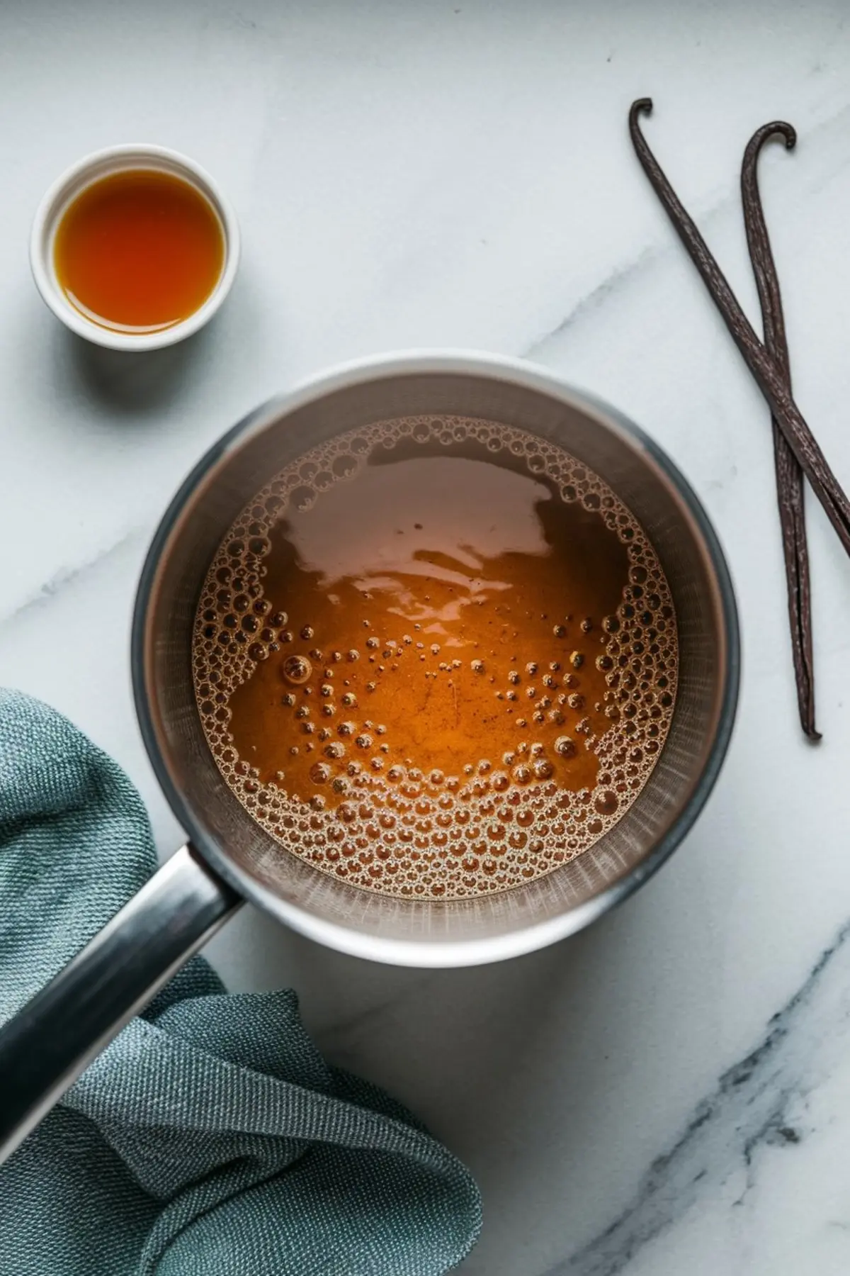 Simmering arnibal syrup made from brown sugar in a stainless steel saucepan, with vanilla beans and a small bowl of syrup on a marble counter, styled with a soft teal cloth.