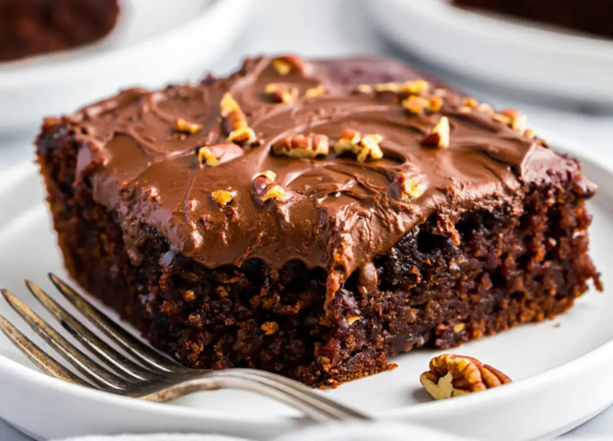 A close-up of a single slice of Texas sheet cake on a white plate with a fork, showing a thick layer of chocolate frosting and chopped pecans on a moist, chocolate-rich base.