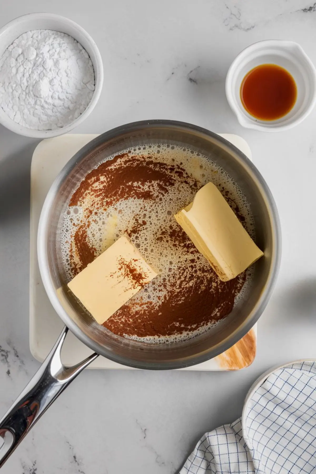 A saucepan containing melted butter, cocoa powder, and sugar on a marble surface, with powdered sugar and vanilla extract nearby, part of the process for making chocolate frosting.