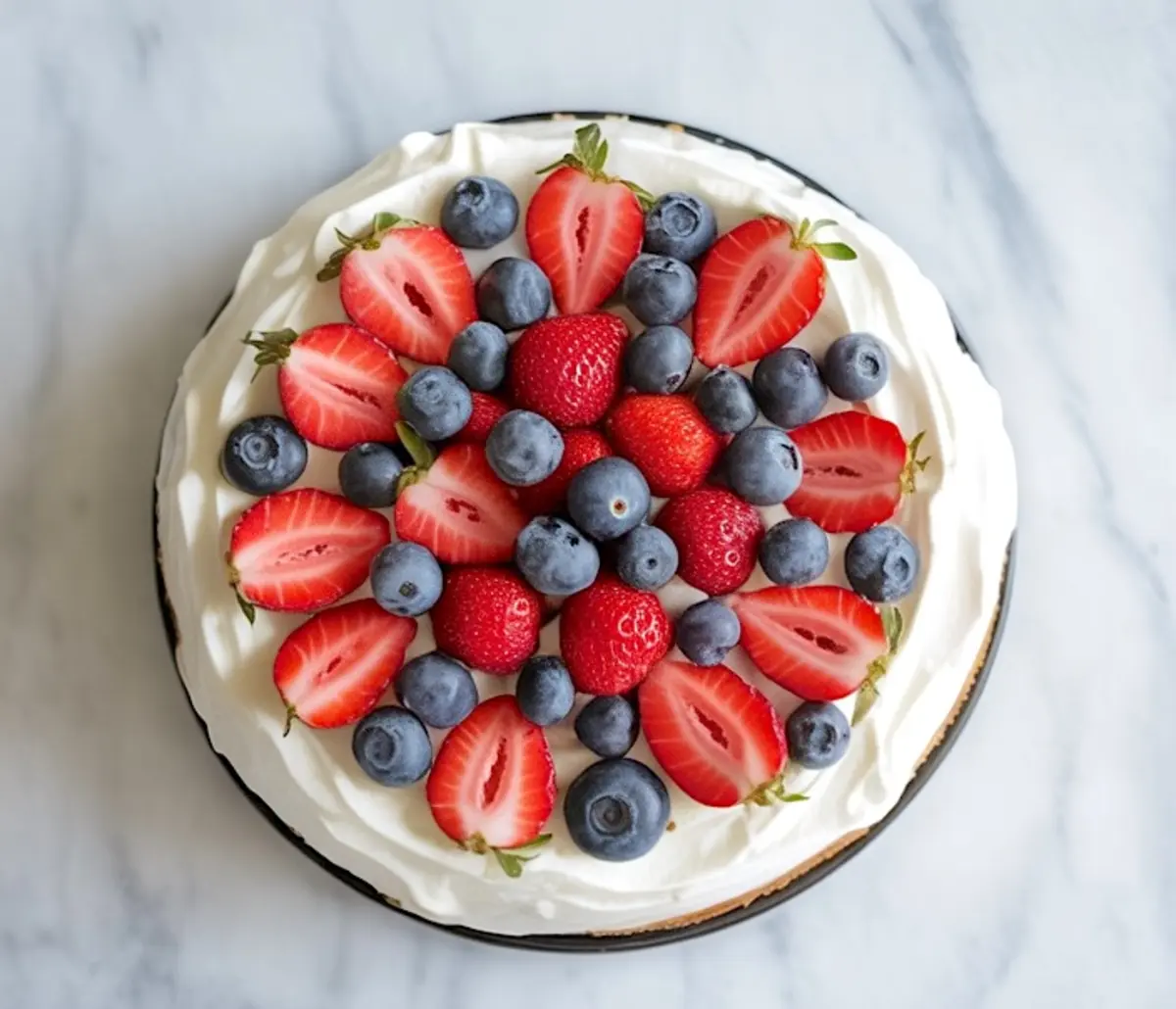 Overhead view of a no-bake berry cheesecake topped with halved strawberries and whole blueberries arranged in a circular pattern over whipped cream on a marble background.