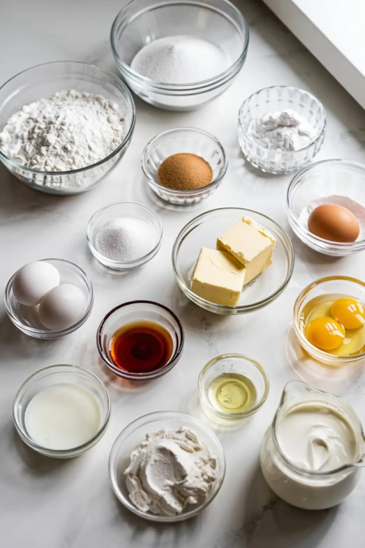 Flat lay of measured vanilla cake ingredients in clear glass bowls, including flour, sugar, butter, eggs, vanilla extract, sour cream, and baking powder on a white countertop.
