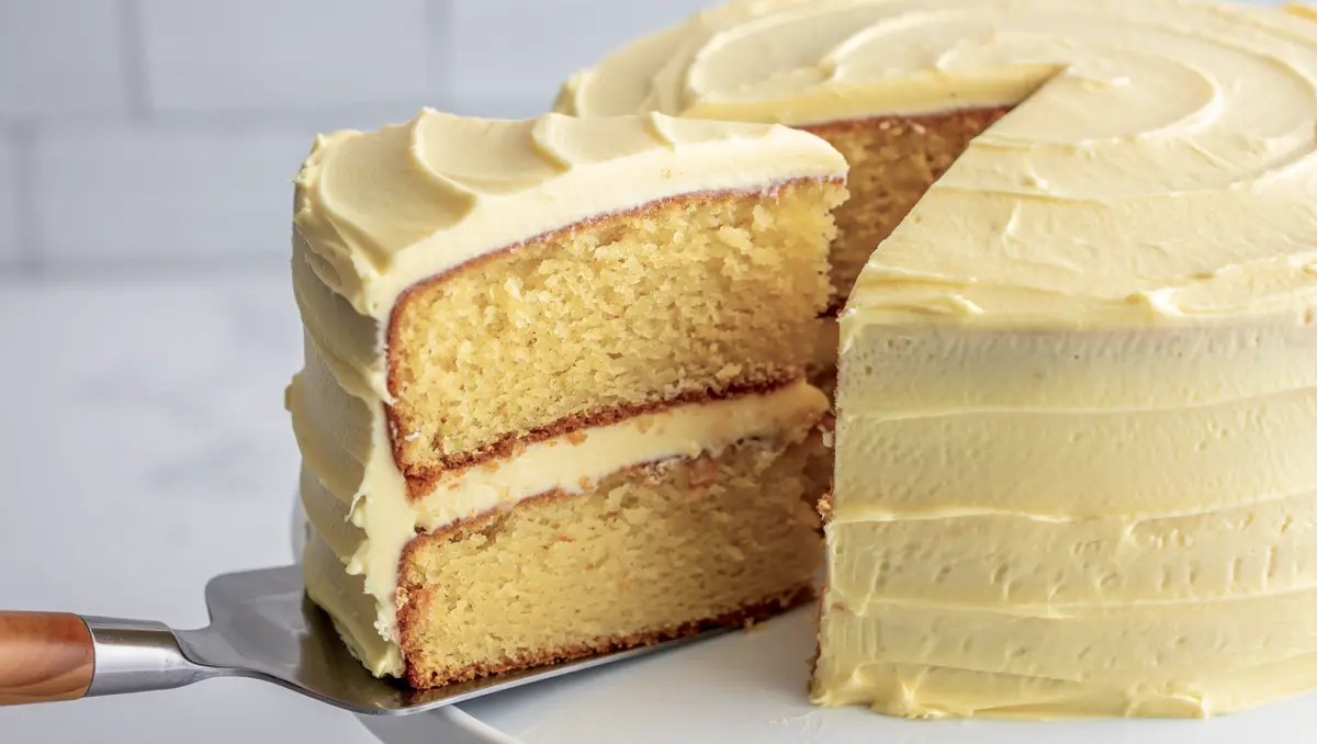 Close-up of a moist vanilla layer cake with yellow buttercream, showcasing the fluffy interior and thick frosting layers as a slice is lifted with a cake spatula.