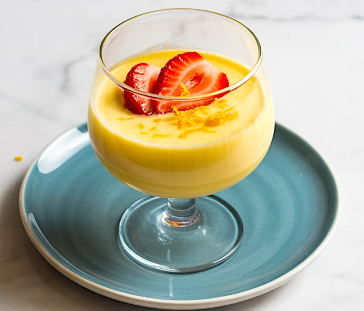 Glass dish of zabaglione served on a teal plate, decorated with halved strawberries and shredded lemon zest, presenting a bright, summery Italian custard dessert on a marble surface.