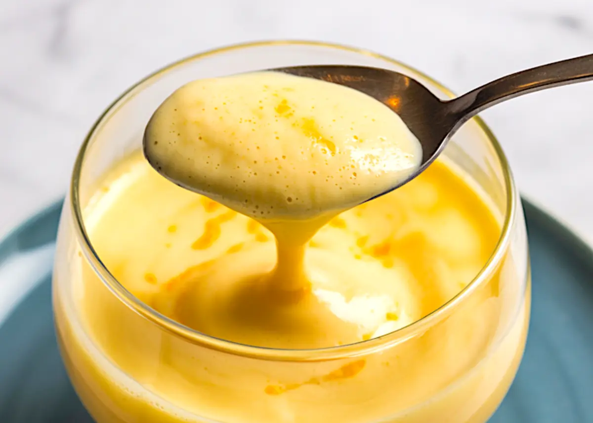 Macro shot of a spoonful of zabaglione above the glass, highlighting the glossy, velvety texture and golden yellow hue of the classic Italian dessert on a marble tabletop.