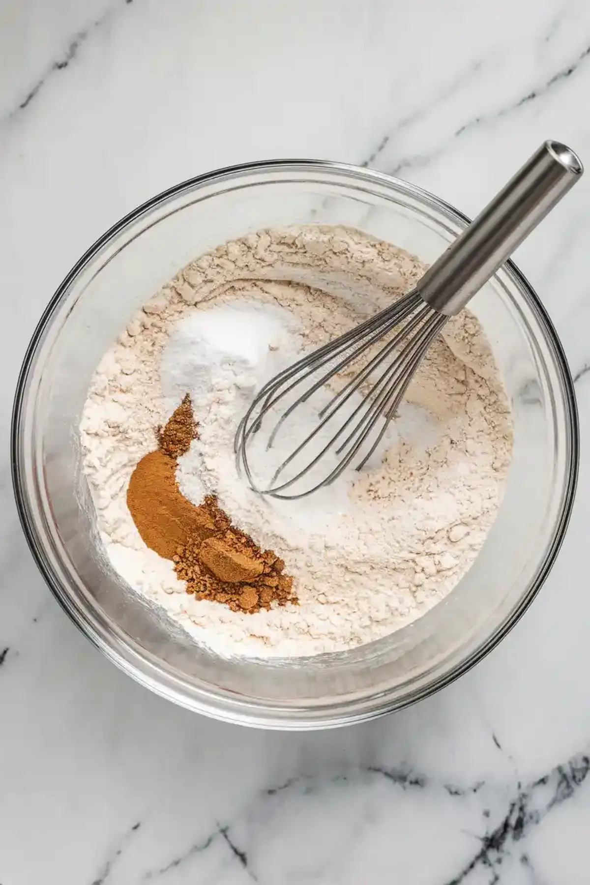 Mixing bowl with flour, baking soda, cinnamon, and nutmeg with a whisk on a marble surface, showing the dry ingredients for banana cake.