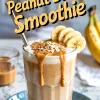 Glass of banana peanut butter smoothie topped with oats, sliced bananas, and drizzled peanut butter, served with a metal straw on a blue plate, with bananas and peanut butter in the background.