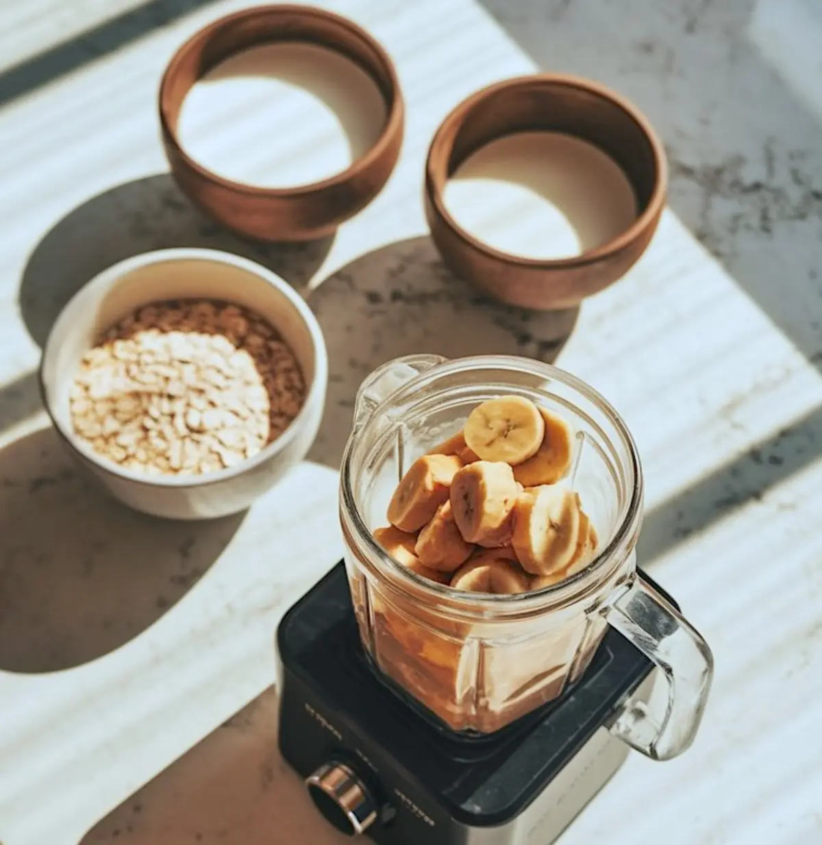 Glass blender filled with sliced bananas and a light brown liquid, set on a blender base beside bowls of oats and plant-based milk on a sunlit countertop.