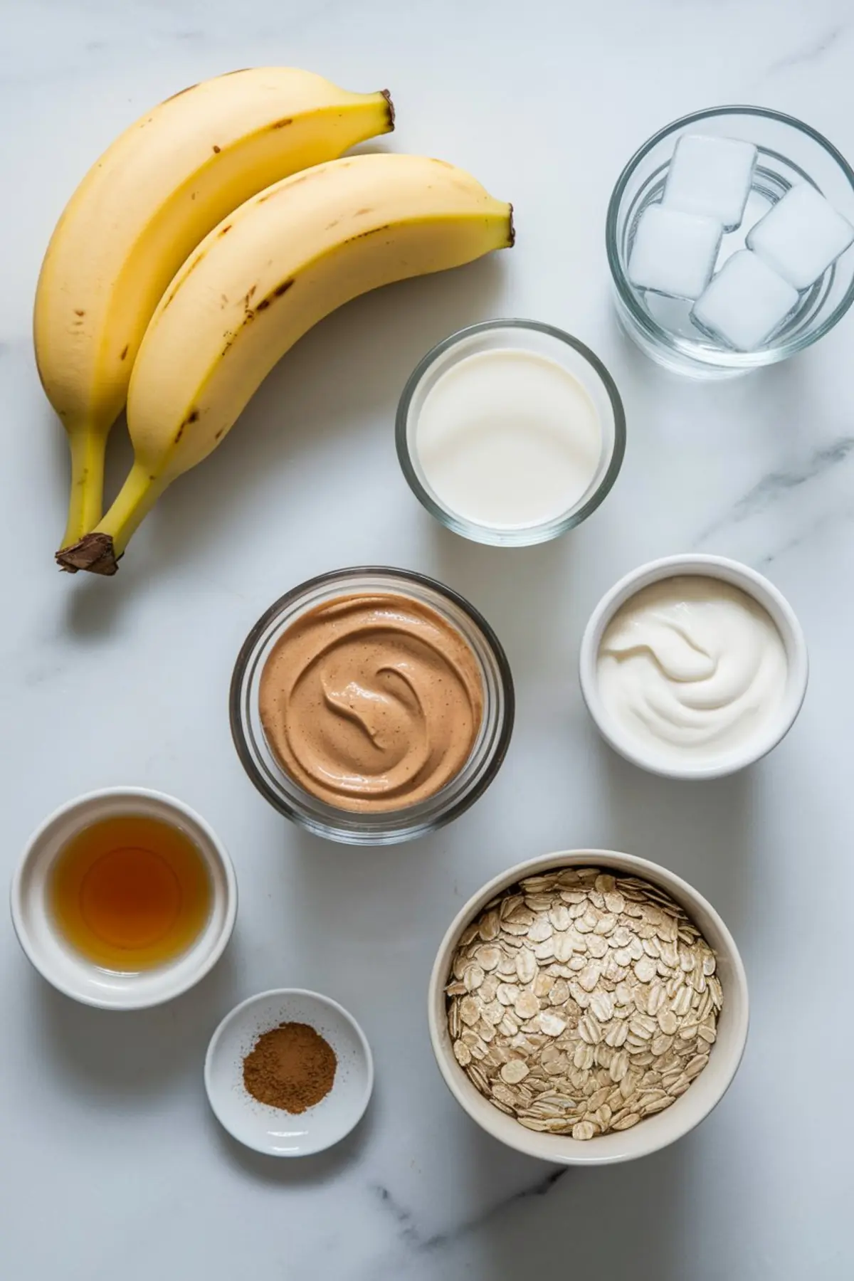 Flat lay of smoothie ingredients on a white surface, including bananas, rolled oats, peanut butter, plant milk, yogurt, vanilla extract, cinnamon powder, and ice cubes in glass bowls.