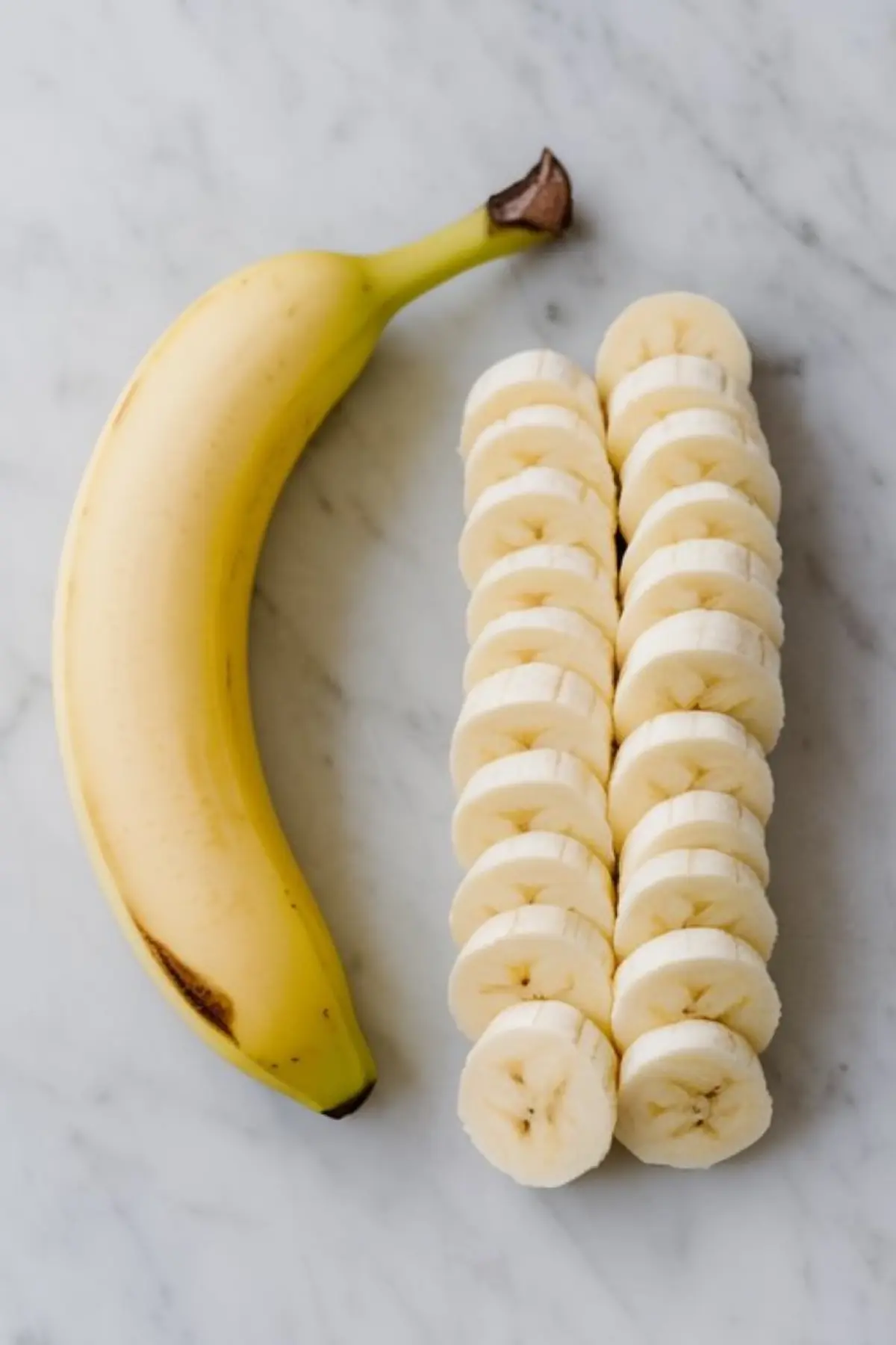A whole ripe banana placed beside evenly sliced banana rounds on a marble surface, showcasing ingredients for desserts or smoothies.