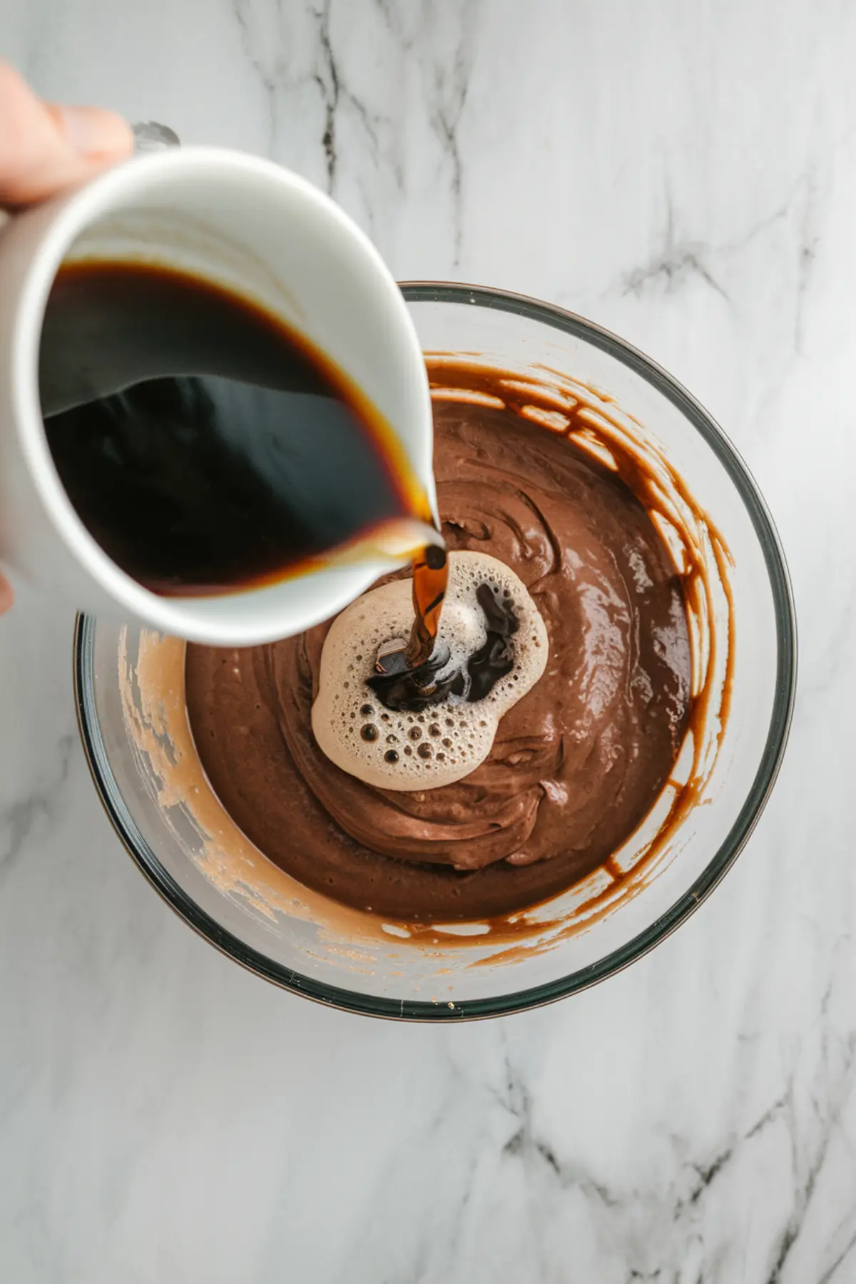 Hot brewed coffee is being poured into a glass bowl of rich chocolate cake batter, capturing the process of enhancing chocolate flavor during baking.