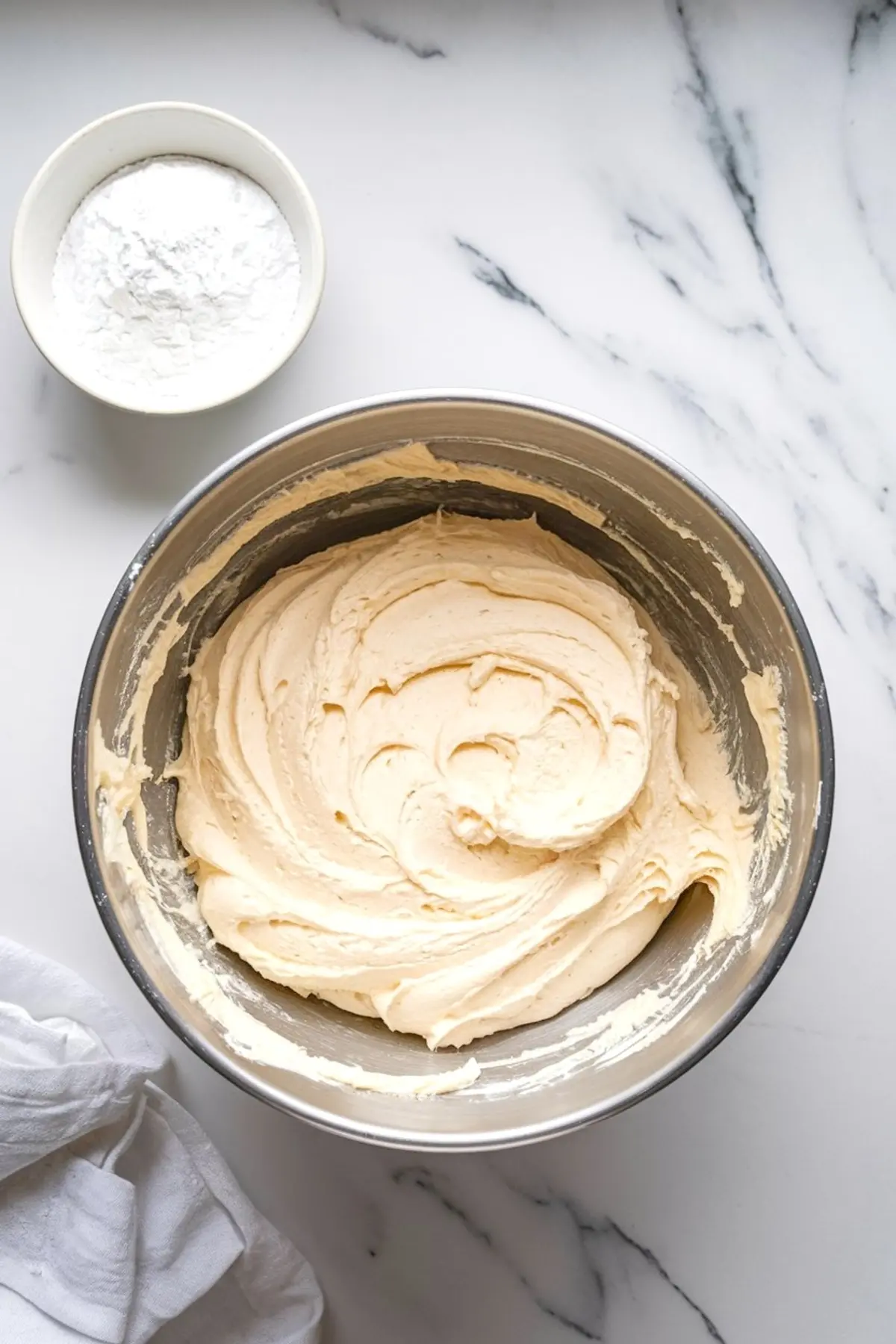 A metal mixing bowl filled with creamy vanilla cake batter sits on a marble countertop next to a small bowl of powdered sugar, showcasing a baking preparation scene.