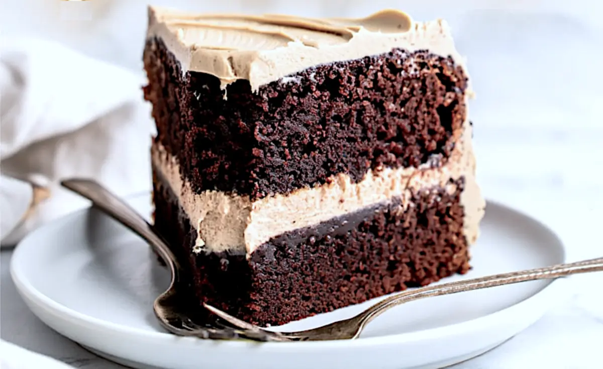 A slice of double-layered chocolate cake with mocha frosting is served on a white plate with a fork, revealing moist texture and creamy filling.