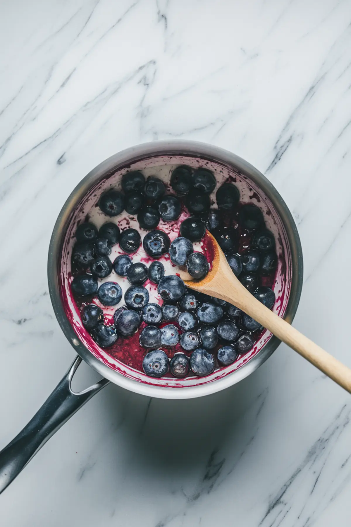 Fresh blueberries being cooked into compote in a saucepan with a wooden spoon, showing dark purple syrup and softened berries on a marble counter.