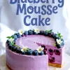 Blueberry mousse cake with a slice removed, showcasing a creamy purple mousse layer filled with whole blueberries, topped with fresh blueberries and white flowers on a golden crust.