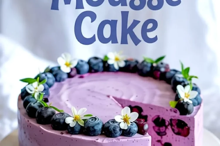 Blueberry mousse cake with a slice removed, showcasing a creamy purple mousse layer filled with whole blueberries, topped with fresh blueberries and white flowers on a golden crust.