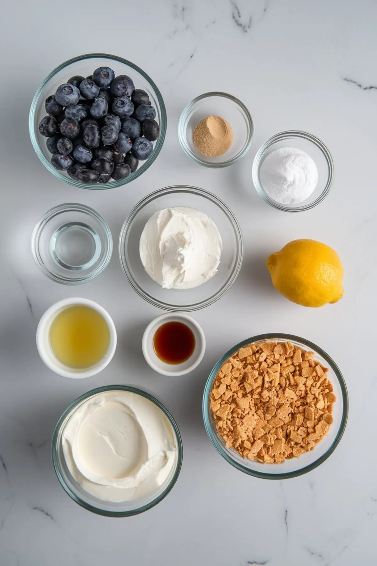 Flat lay of ingredients for a blueberry mousse cake including fresh blueberries, cream cheese, lemon, gelatin, sour cream, vanilla, lemon juice, sugar, and crushed cookies in glass bowls.