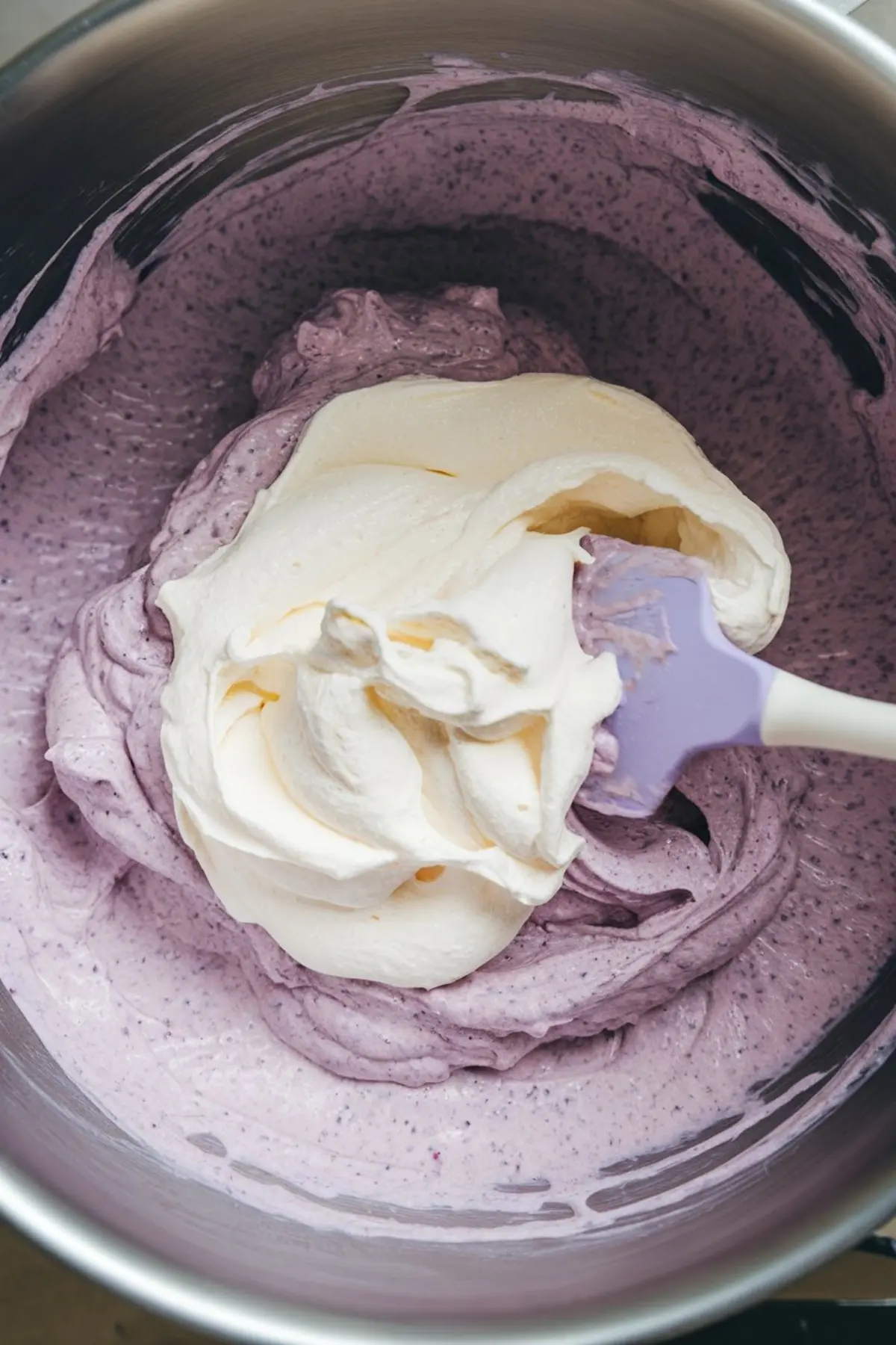 Purple blueberry mousse being folded with whipped cream in a mixing bowl using a silicone spatula, creating a light and airy filling.