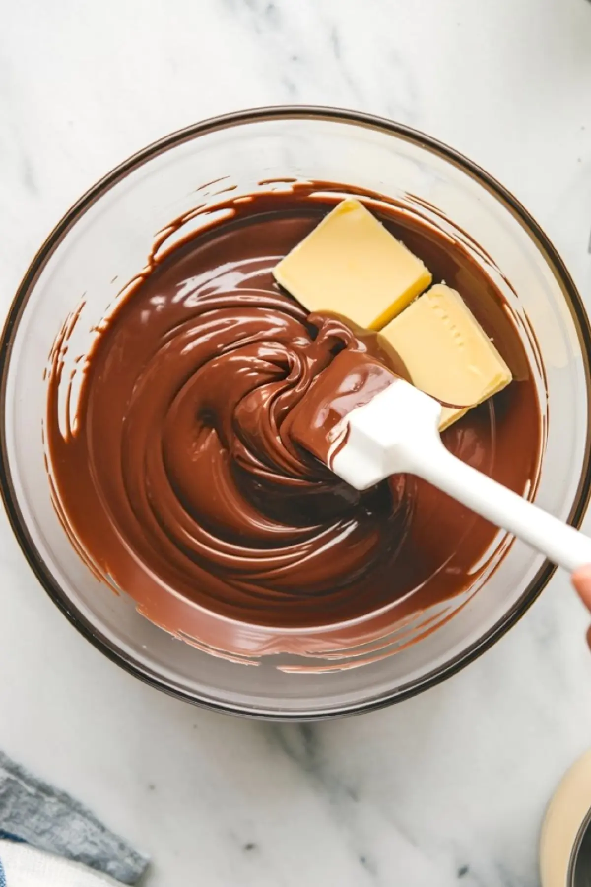 Glossy melted chocolate folds around butter cubes with a white spatula in a glass bowl, illustrating the ganache base for fudgy brownie cookies.