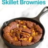 Baked caramel apple brownie in a cast iron skillet, topped with caramel drizzle and golden apple chunks, set on a white marble background with clear text overlay.
