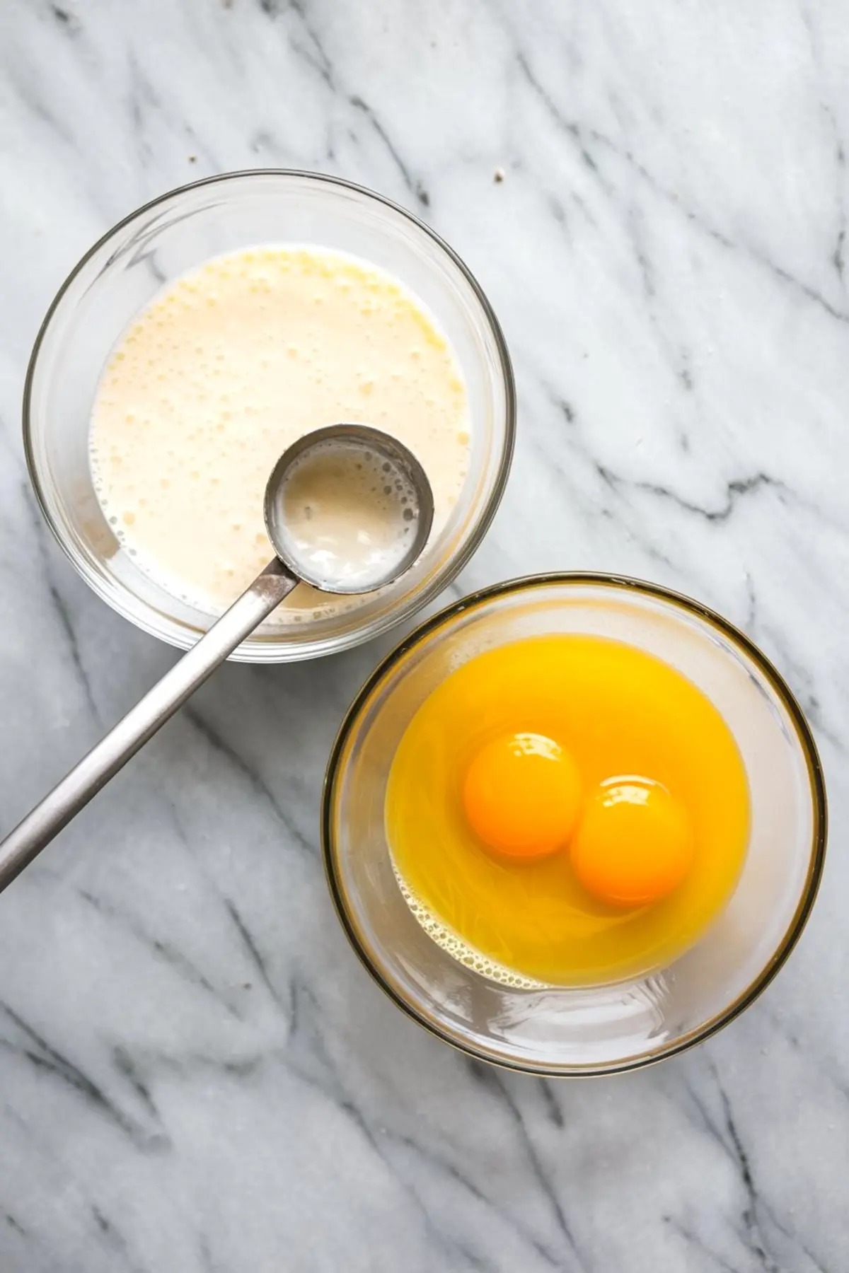 Two glass bowls on a marble counter, one with cracked eggs and one with a creamy liquid mixture, with a metal ladle resting inside.