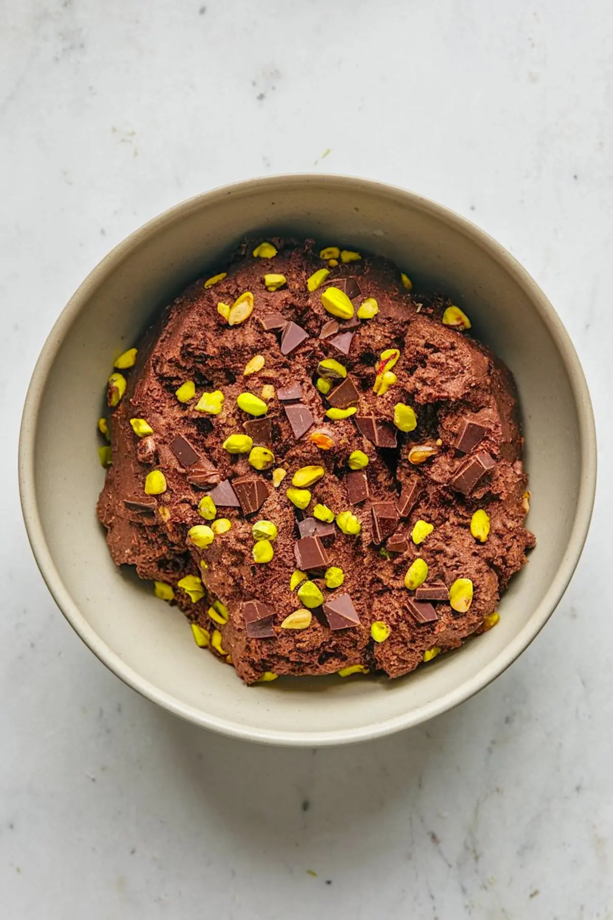 Bowl of chocolate biscotti dough mixed with chopped dark chocolate and bright green pistachios. The dough has a rich, dense texture ready for baking.