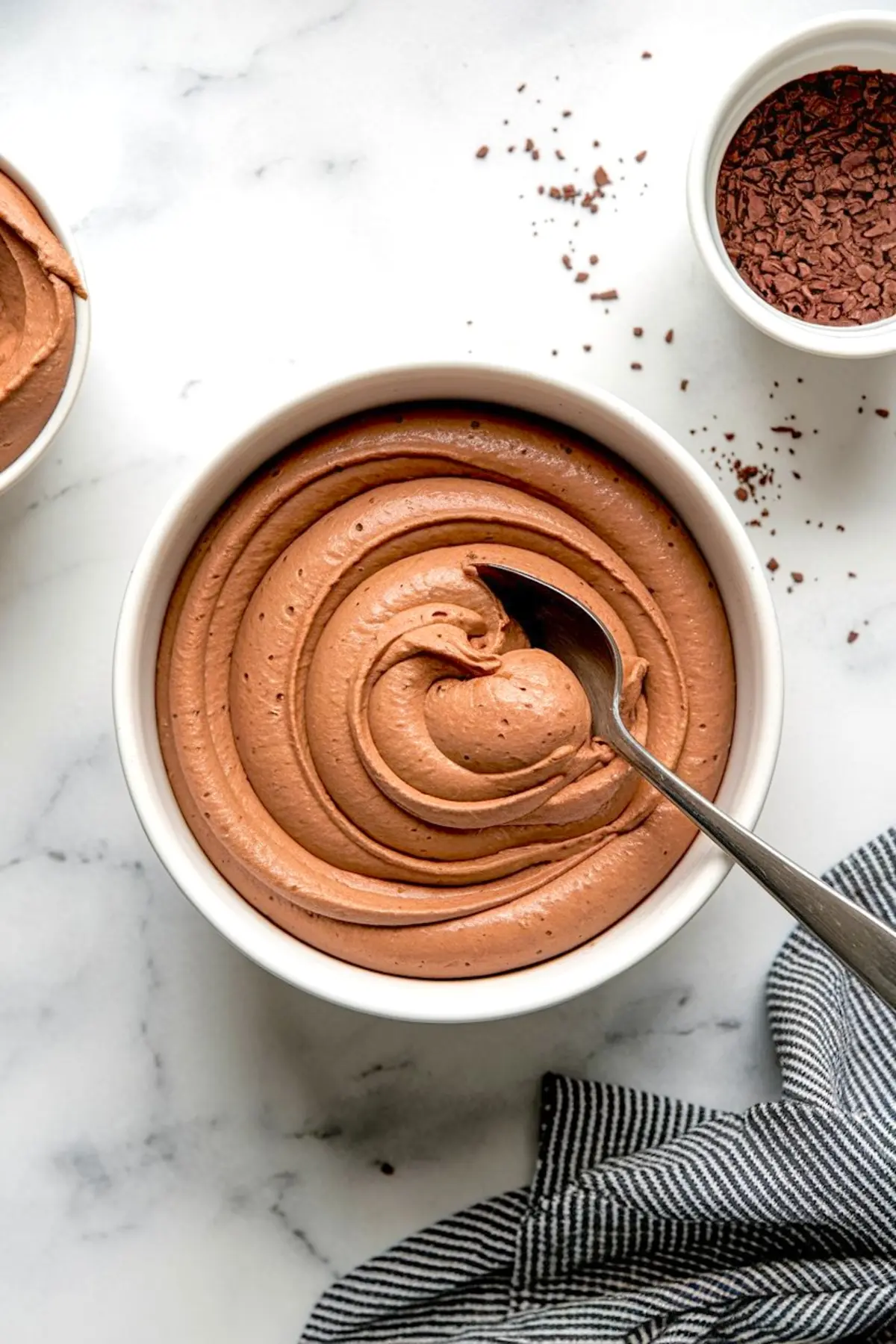Smooth chocolate mousse filling swirled in a white bowl with a spoon, ready for assembling the chocolate raspberry cheesecake domes, on a marble countertop with cocoa flakes scattered around.
