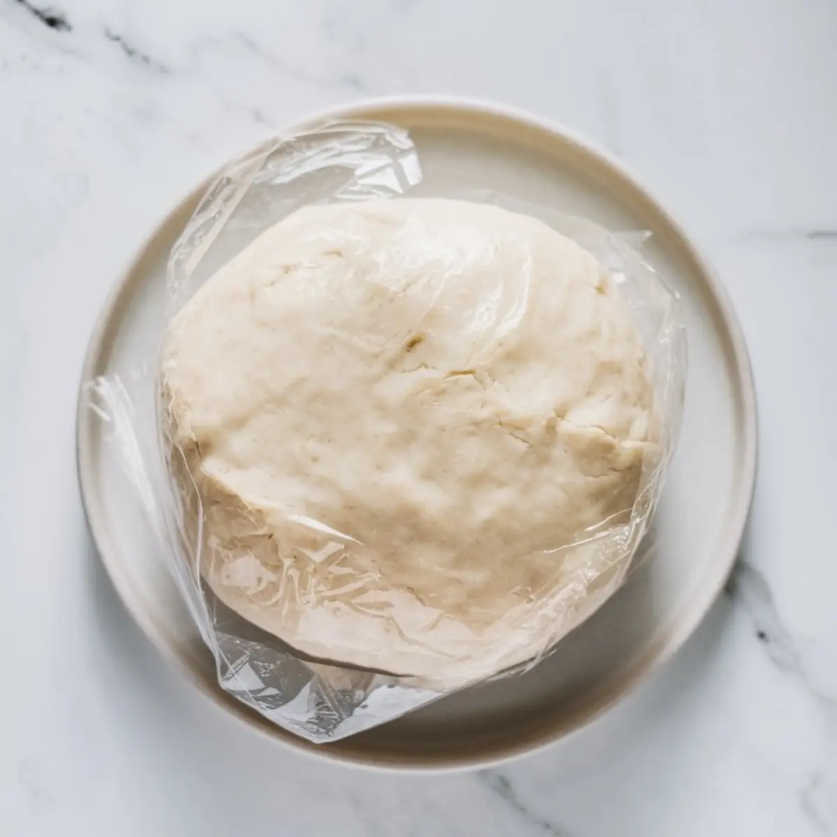 Ball of cream cheese cookie dough wrapped in plastic on a white plate. The dough has a smooth, soft texture, resting on a marble countertop.
