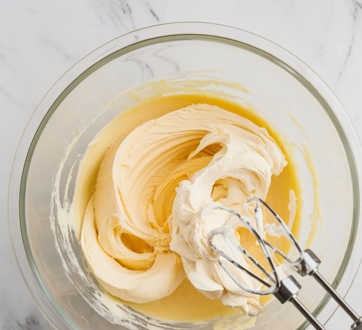 Cream cheese cookie batter in a glass mixing bowl with electric mixer beaters. The mixture is light and creamy with a smooth, fluffy texture.
