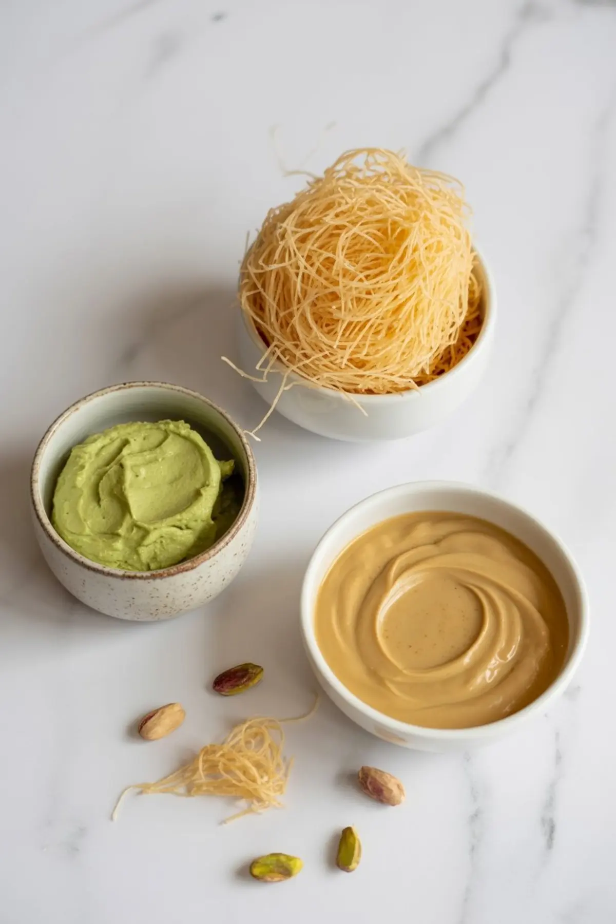 Three small bowls on a white surface containing raw golden kataifi pastry, green pistachio cream, and smooth caramel-like spread, with pistachios and vermicelli strands scattered around for a styled ingredient shot.