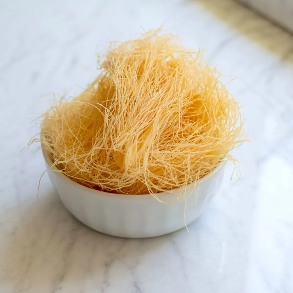 A white ramekin filled with uncooked golden kataifi pastry strands, placed on a marble surface for a clean and minimal food preparation visual.