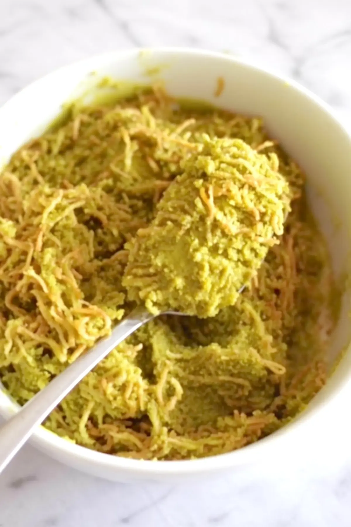 A close-up of a white bowl containing green pistachio chocolate filling mixed with golden vermicelli, with a spoonful lifted to highlight the fibrous, creamy texture.