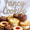 Decorative plate filled with assorted sandwich cookies, including chocolate-dipped, jam-filled, and cream-filled cookies, topped with edible gold flakes and a “Fancy Cookies” sign in the background.