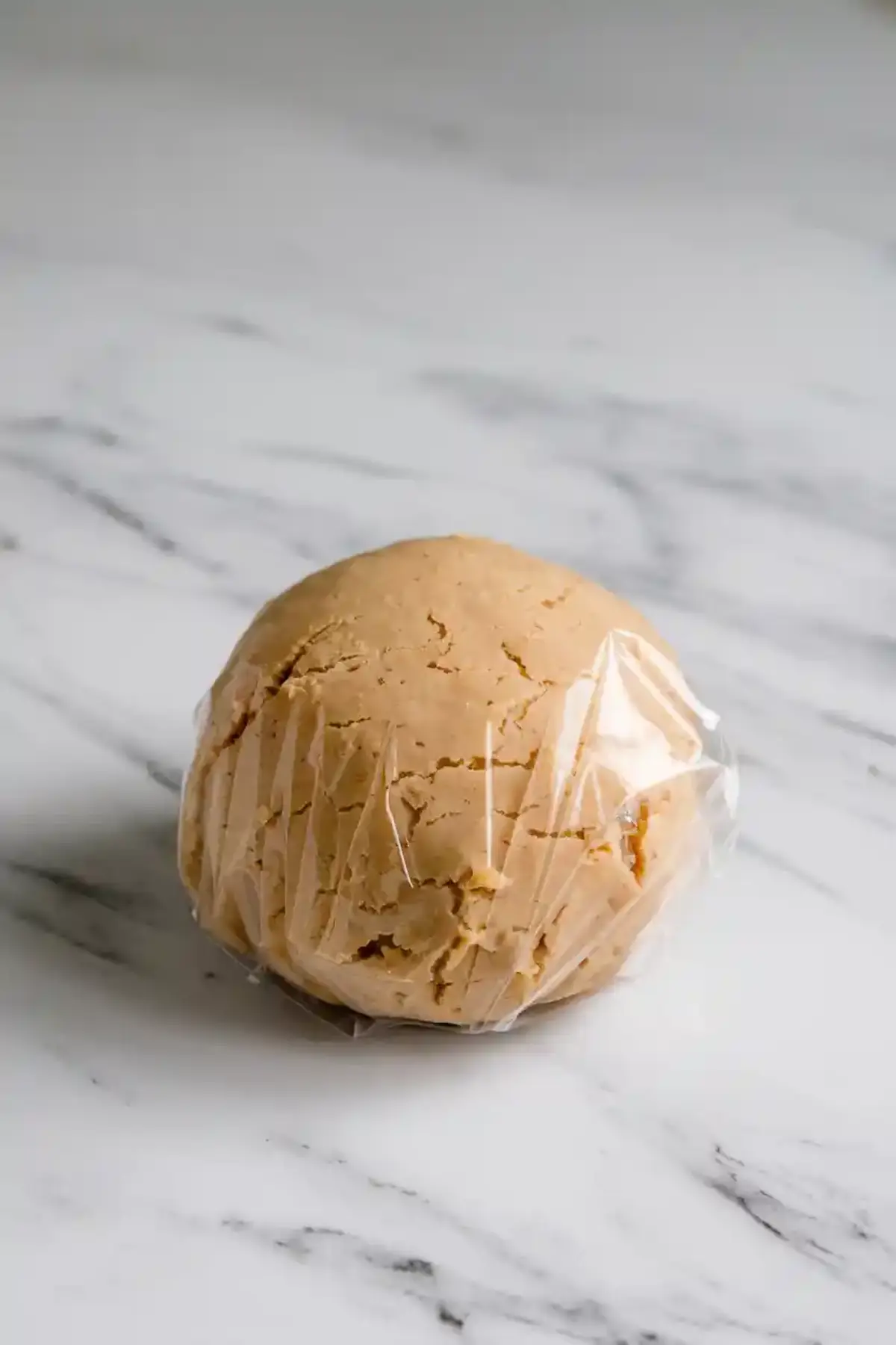  Ball of cookie dough wrapped in plastic wrap resting on a white marble countertop, ready for chilling before baking.