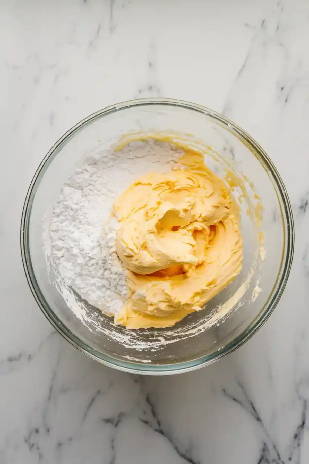 Creamy butter and powdered sugar mixture partially blended in a glass bowl on a marble surface, showing the start of cookie dough preparation.