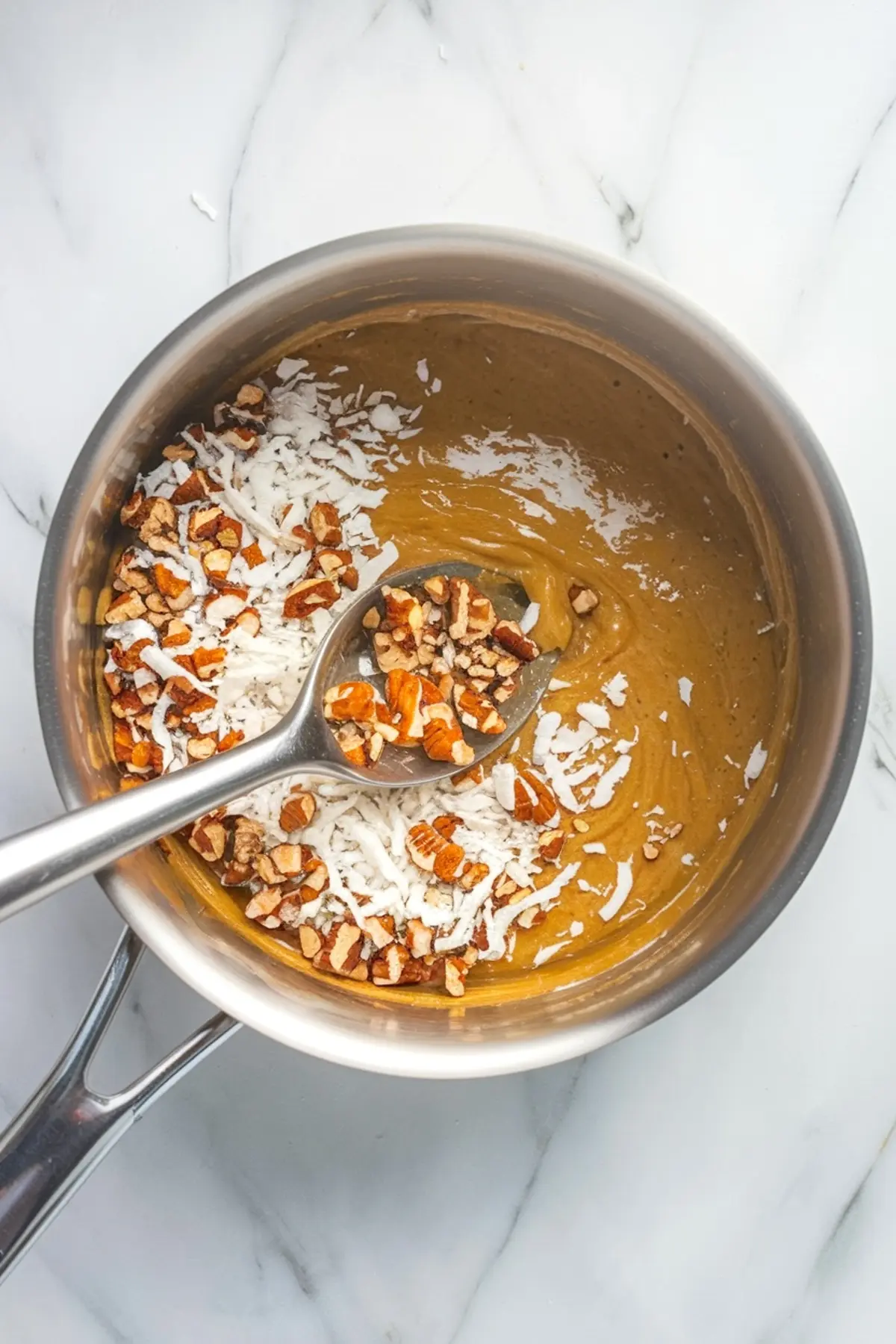 Saucepan containing coconut pecan filling with shredded coconut and chopped pecans being stirred into a golden caramel base.