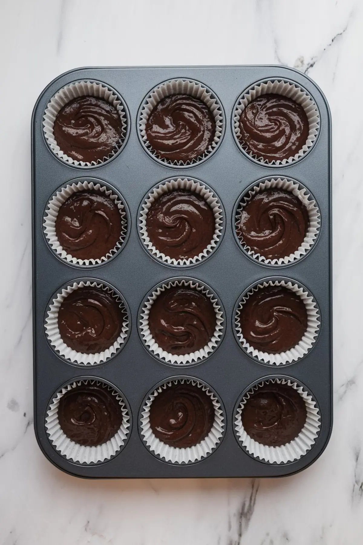 Overhead view of a muffin tin filled with swirled chocolate cupcake batter in white paper liners on a marble surface.